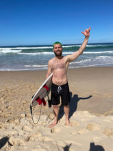 Load image into Gallery viewer, Smiling man on beach wearing Salty Johnson sustainable mens board shorts holding surfboard under arm