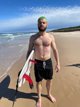 Load image into Gallery viewer, Man on beach wearing sustainable mens board shorts by Salty Johnson holding a surfboard