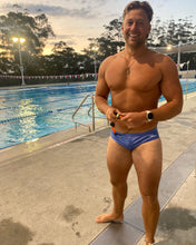 Load image into Gallery viewer, Man wearing Salty Johnson sustainable mens swim briefs standing by an outdoor swimming pool at sunset