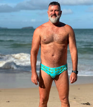 Load image into Gallery viewer, Man wearing blue Sustainable Mens Swim Briefs with tropical print standing on beach by ocean