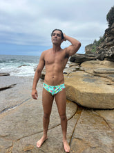 Load image into Gallery viewer, Male model wearing Sustainable Mens Swim Briefs with colorful pattern standing on rocky beach by ocean