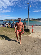 Load image into Gallery viewer, Man wearing Salty Johnson sustainable mens swim briefs in red with white print at the beach on a sunny day - Size 34