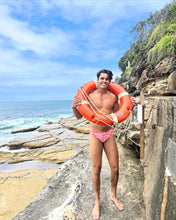 Load image into Gallery viewer, Man wearing Salty Johnson AU sustainable mens swim briefs holding an orange lifesaver by the rocky ocean shore