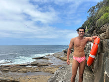 Load image into Gallery viewer, Man wearing Salty Johnson AU sustainable mens swim briefs standing on rocky ocean shore holding a lifebuoy
