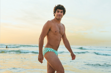 Load image into Gallery viewer, Young man wearing sustainable mens swim briefs by Salty Johnson AU walking on the beach at sunset