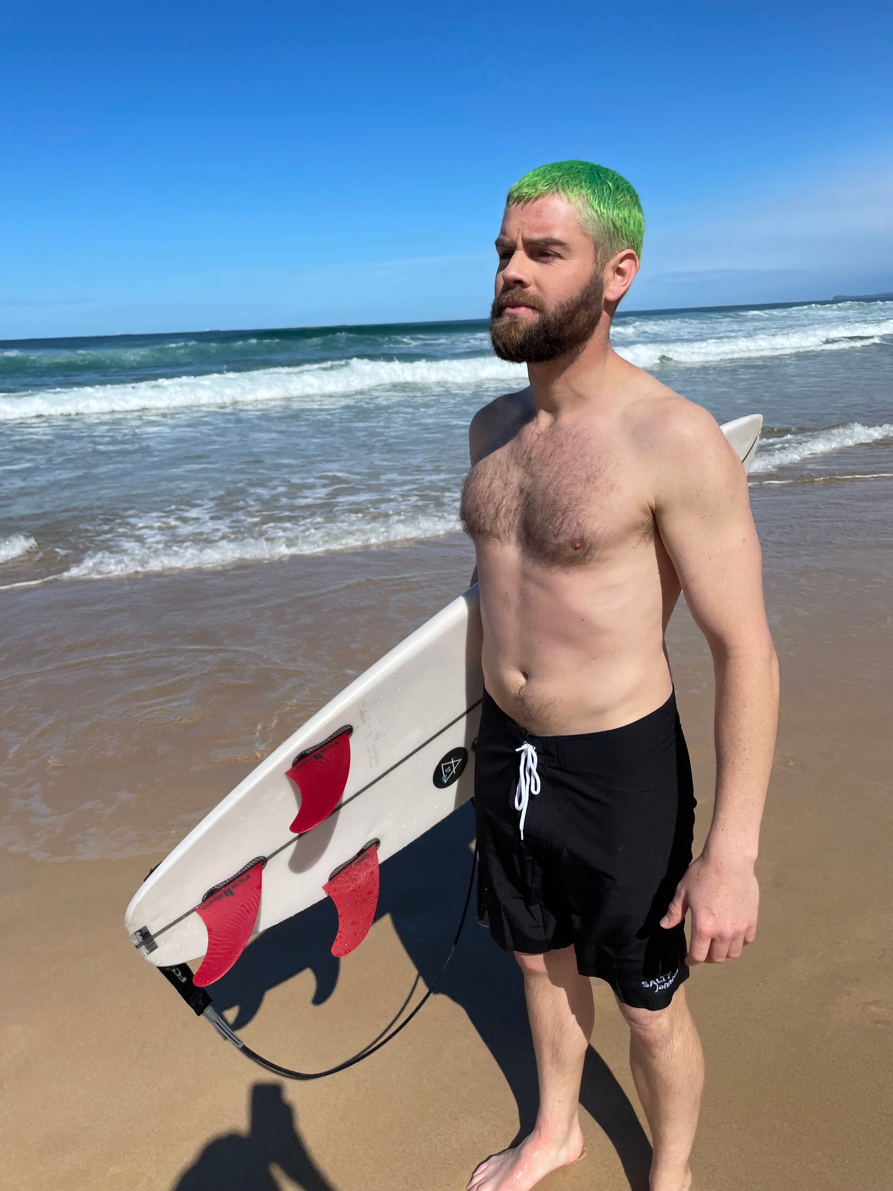 Man with green hair standing on the beach holding a surfboard wearing Salty Johnson sustainable mens board shorts