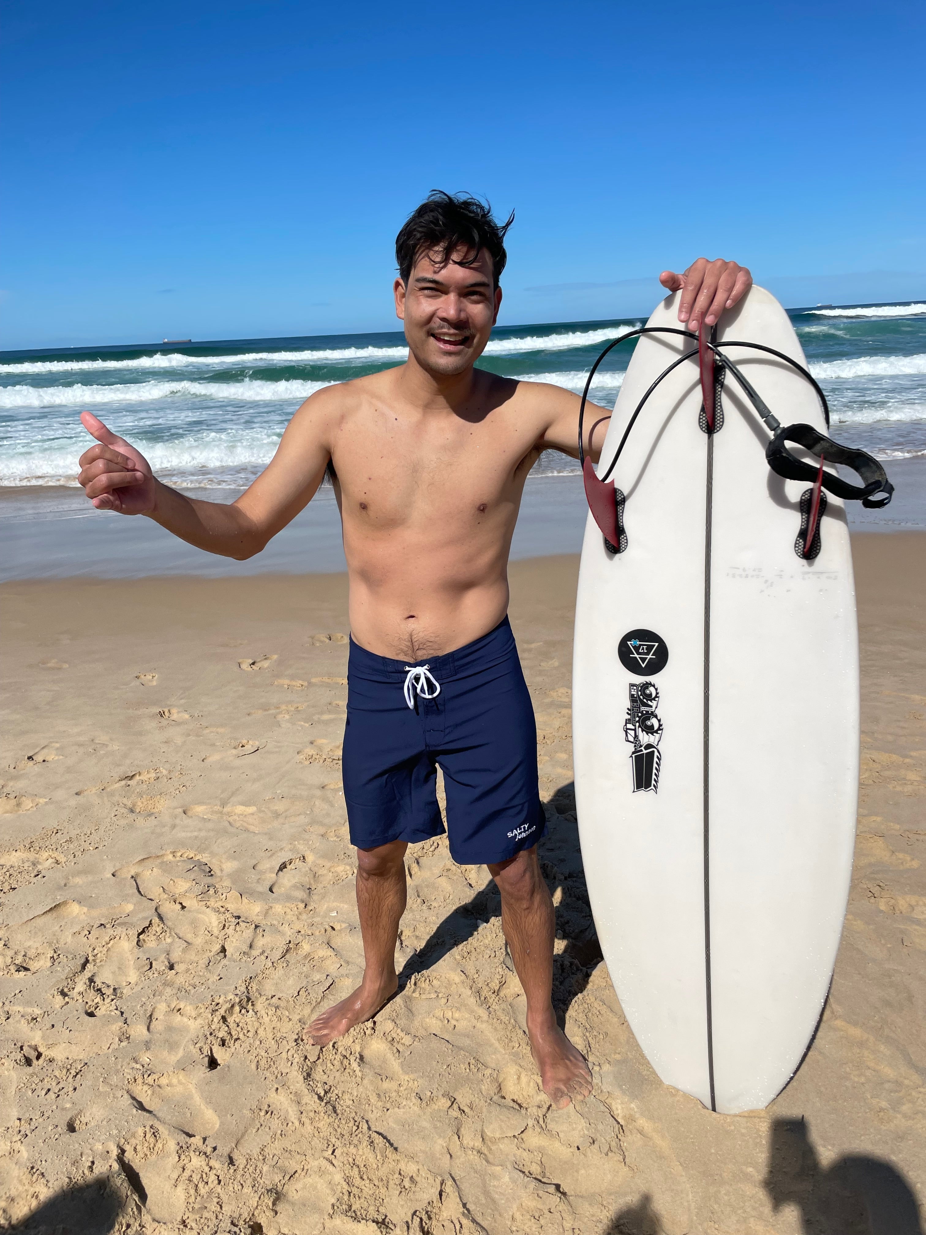 Man on beach wearing Salty Johnson sustainable mens board shorts holding white surfboard