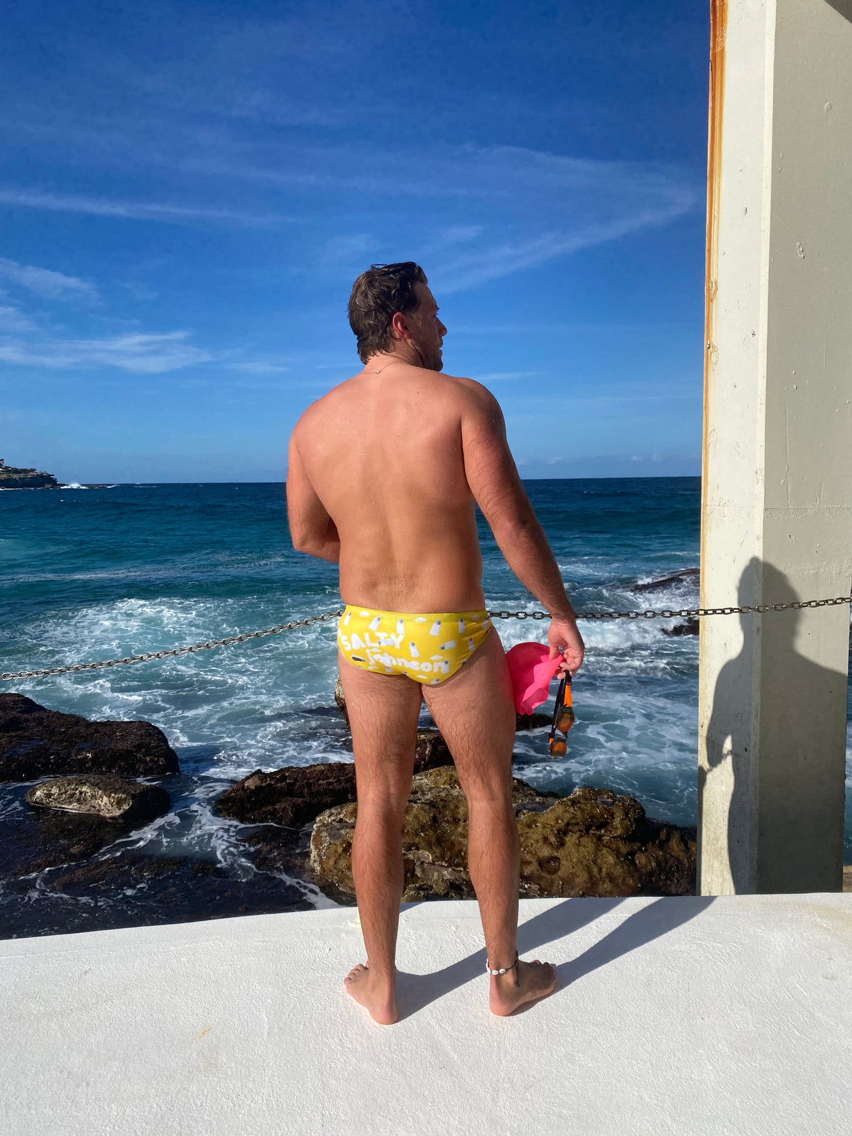 Man standing by ocean wearing yellow sustainable mens swim briefs by Salty Johnson holding pink goggles