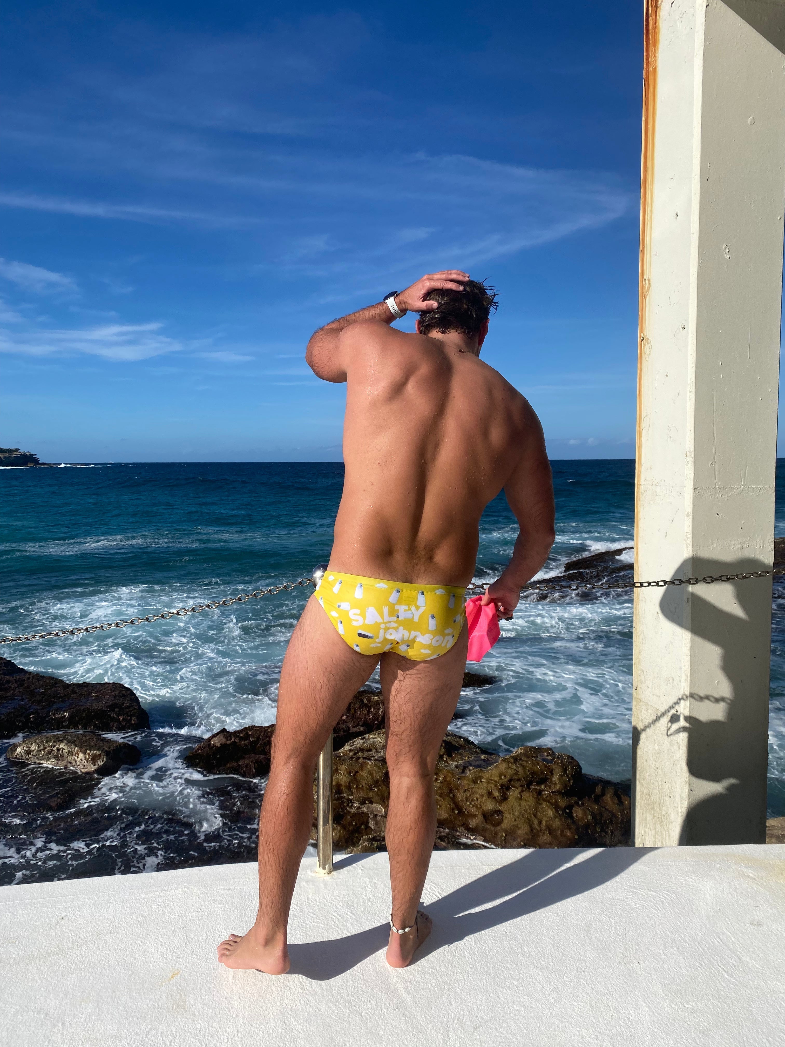 Man wearing yellow Salty Johnson sustainable mens swim briefs standing by ocean with clear blue sky