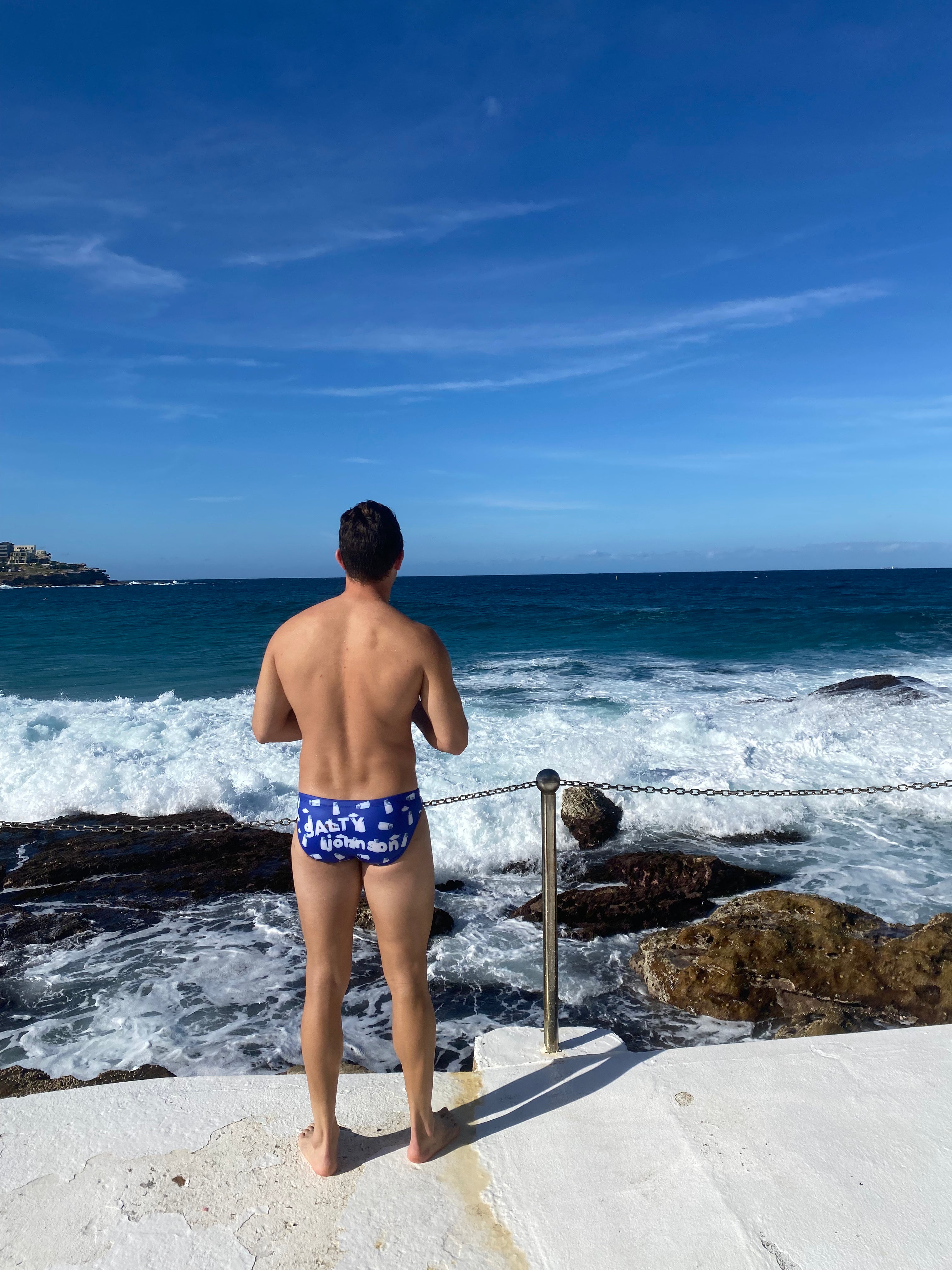 Man wearing Salty Johnson sustainable mens swim briefs standing by rocky ocean shore on a sunny day