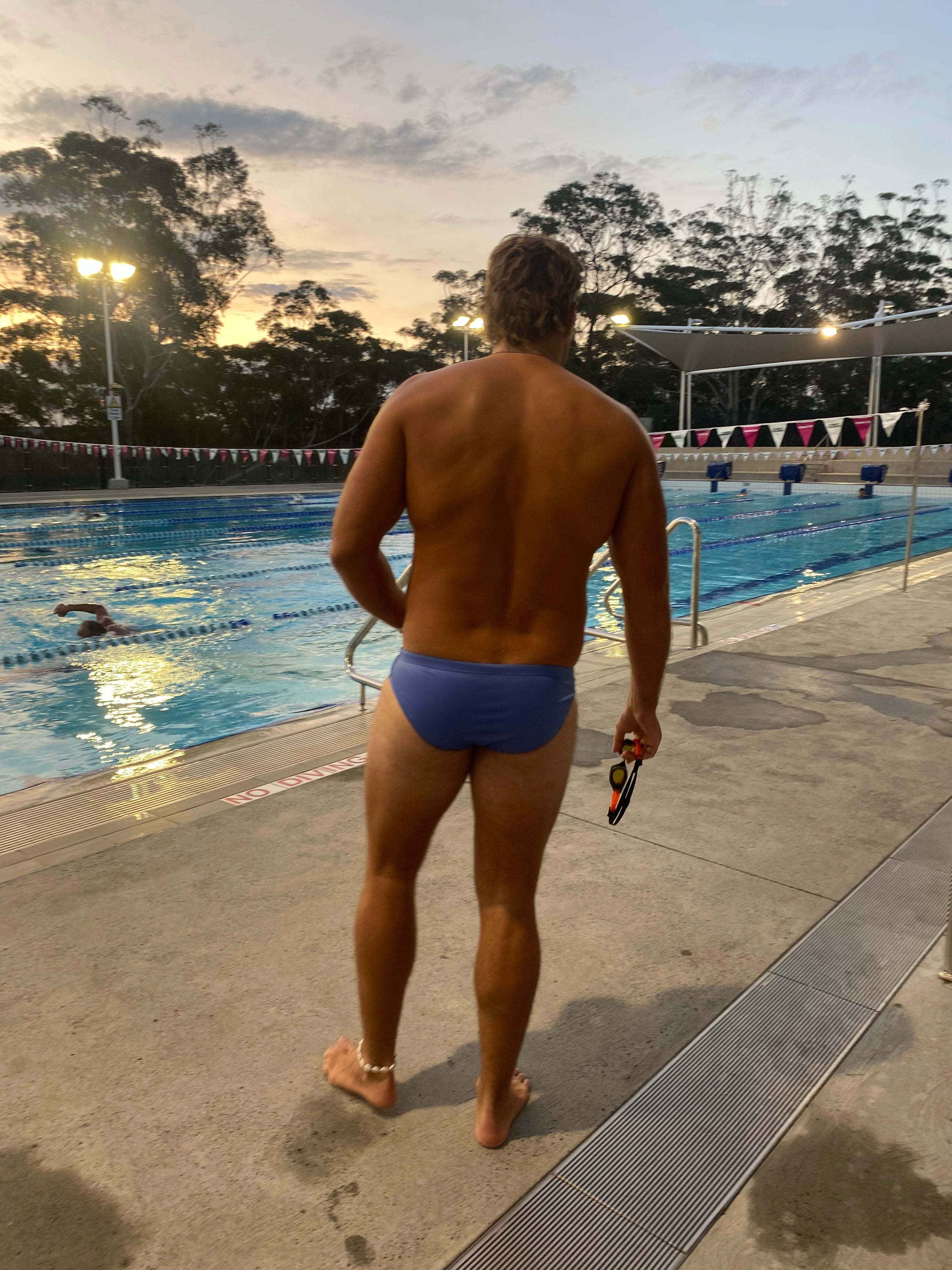 Man wearing Salty Johnson sustainable mens swim briefs standing by outdoor pool at sunset