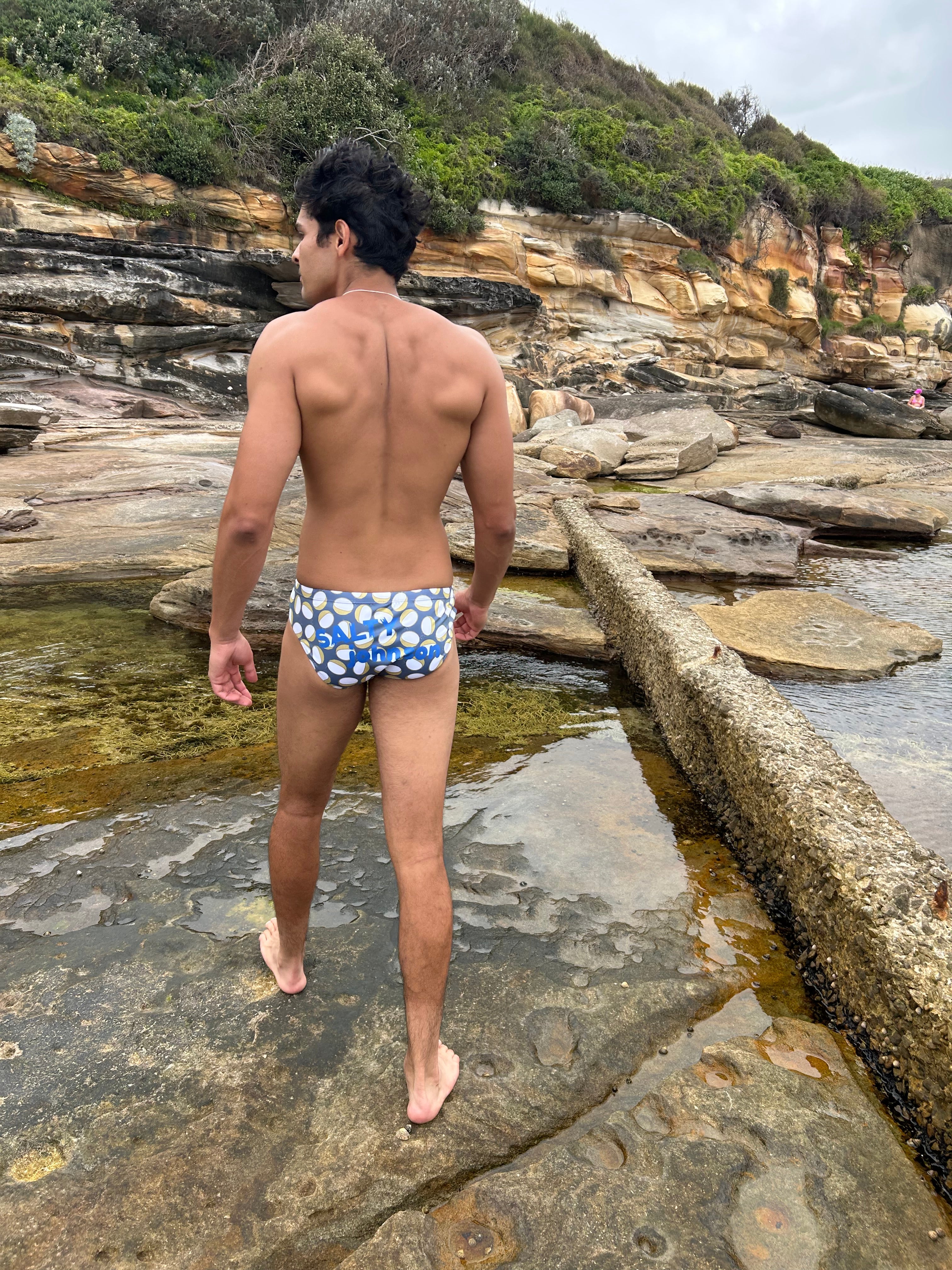 Man wearing Sustainable Mens Swim Briefs by Salty Johnson AU standing on rocky shore near water