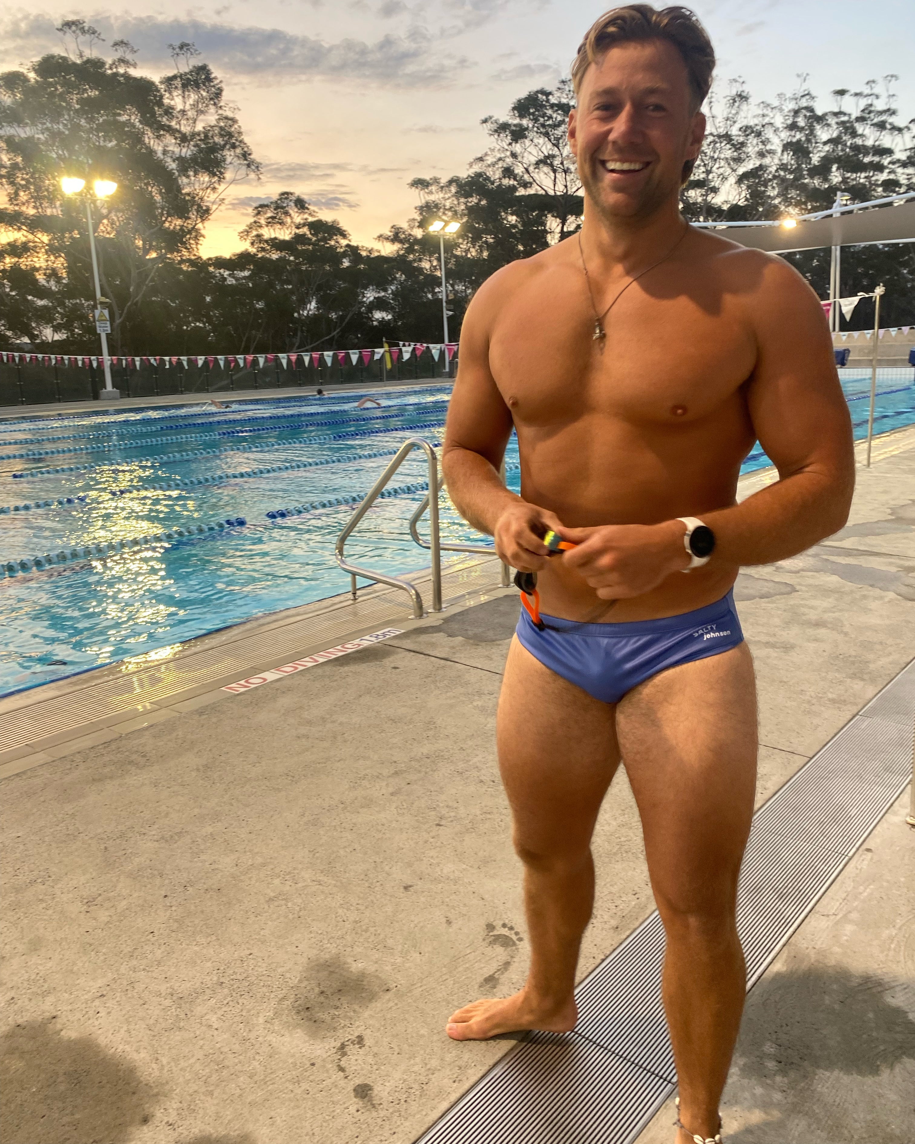 Man wearing Salty Johnson sustainable mens swim briefs standing by an outdoor swimming pool at sunset