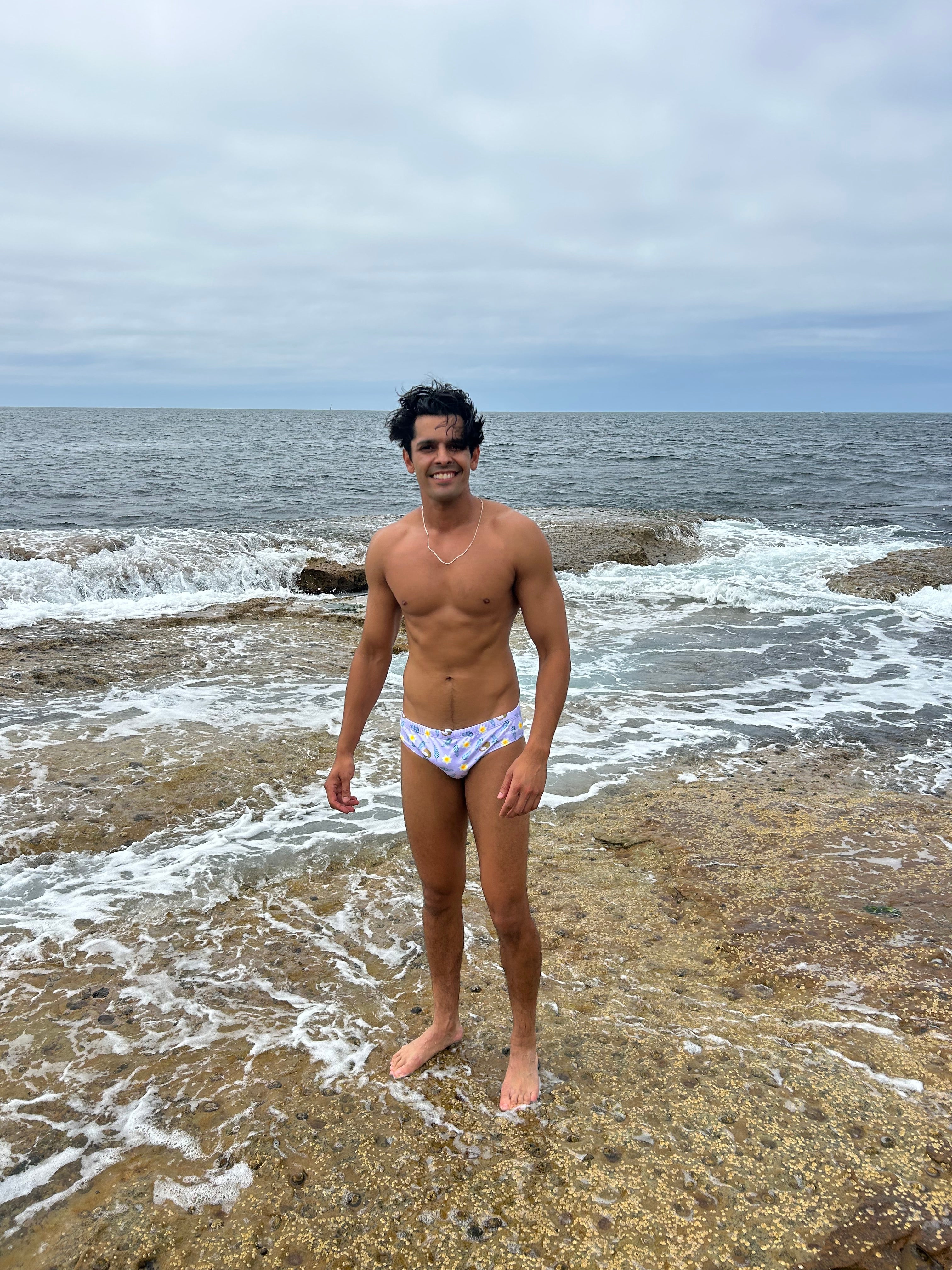 Man wearing Sustainable Mens Swim Briefs by Salty Johnson AU standing on rocky shore near ocean waves