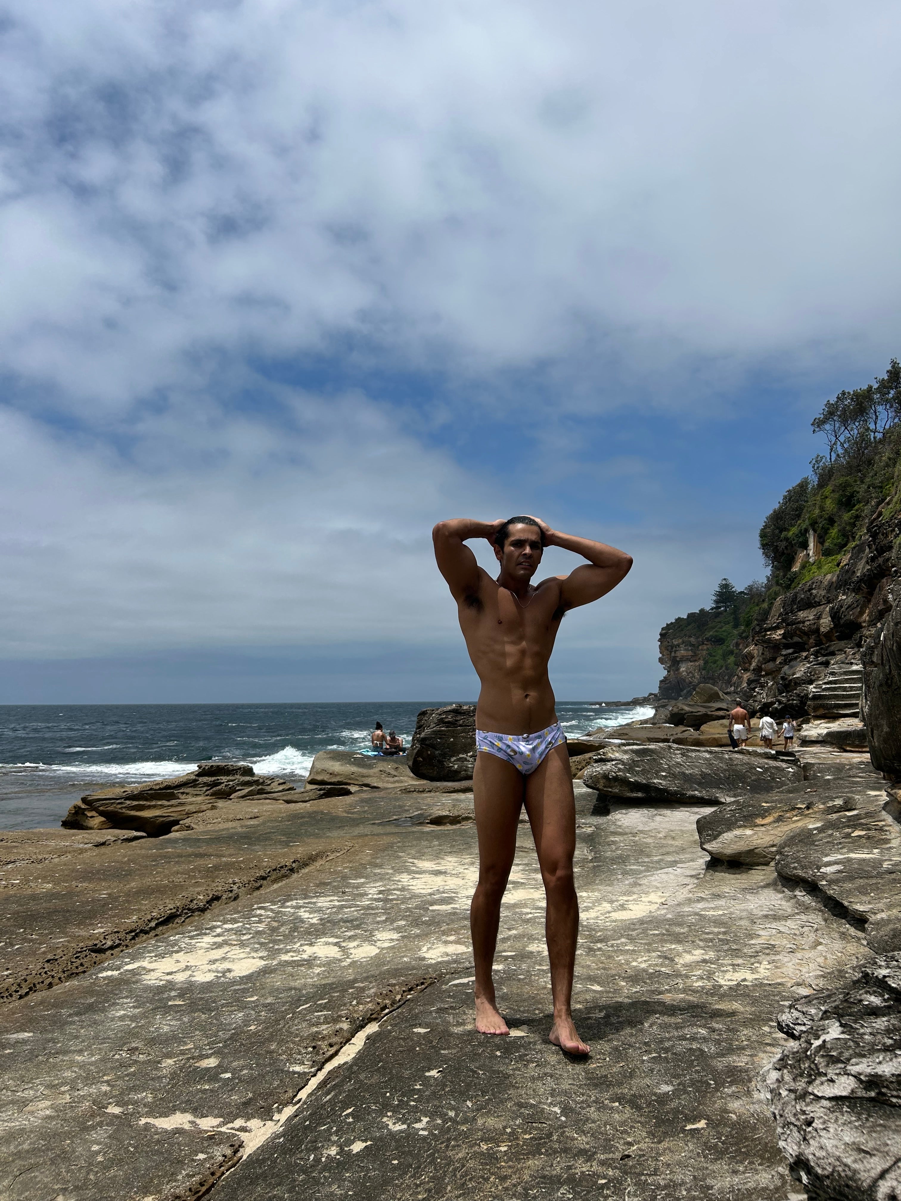 Man wearing Salty Johnson AU sustainable mens swim briefs standing on rocky beach under cloudy sky