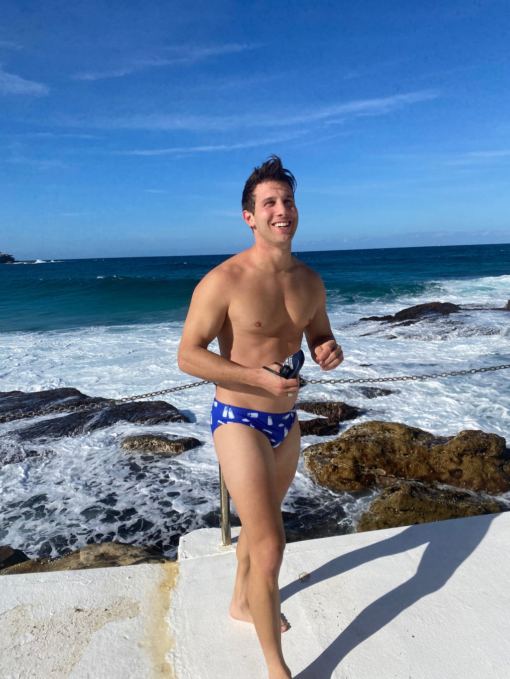 Man wearing Salty Johnson sustainable mens swim briefs at rocky ocean shore under blue sky