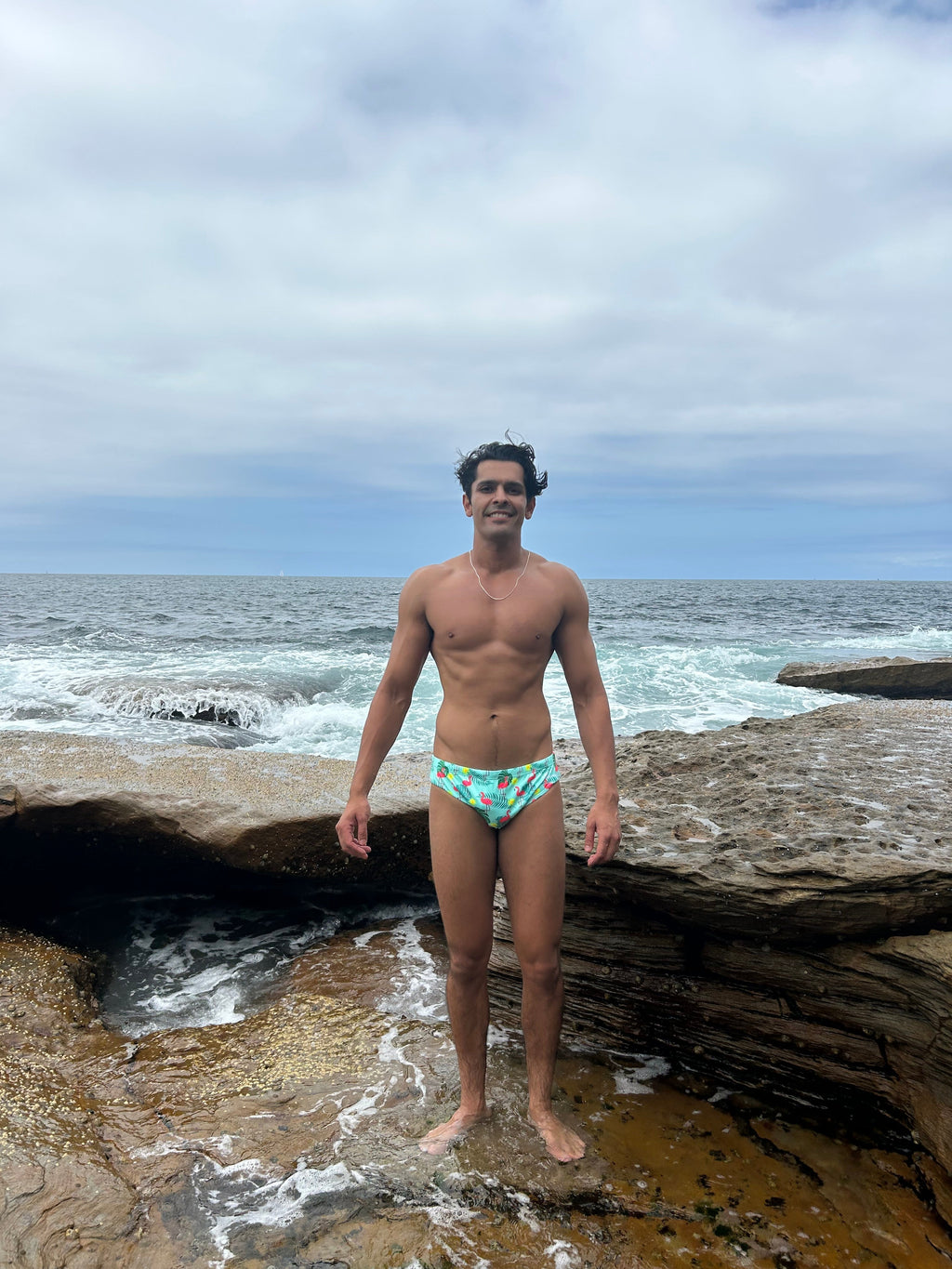 Man wearing sustainable mens swim briefs by Salty Johnson AU standing on rocky shore with ocean waves in background