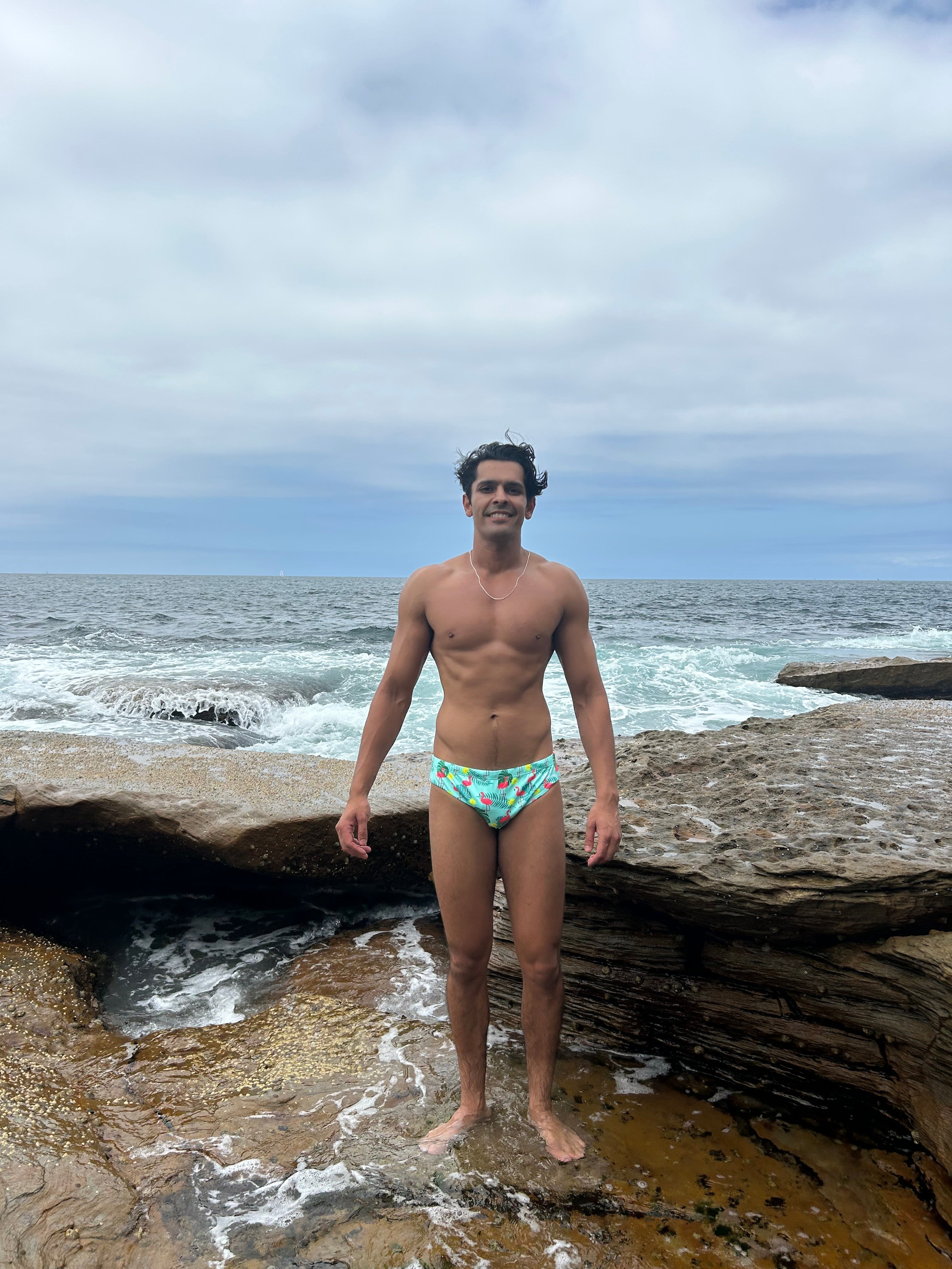 Man wearing sustainable mens swim briefs by Salty Johnson AU standing on rocky shore with ocean waves in background