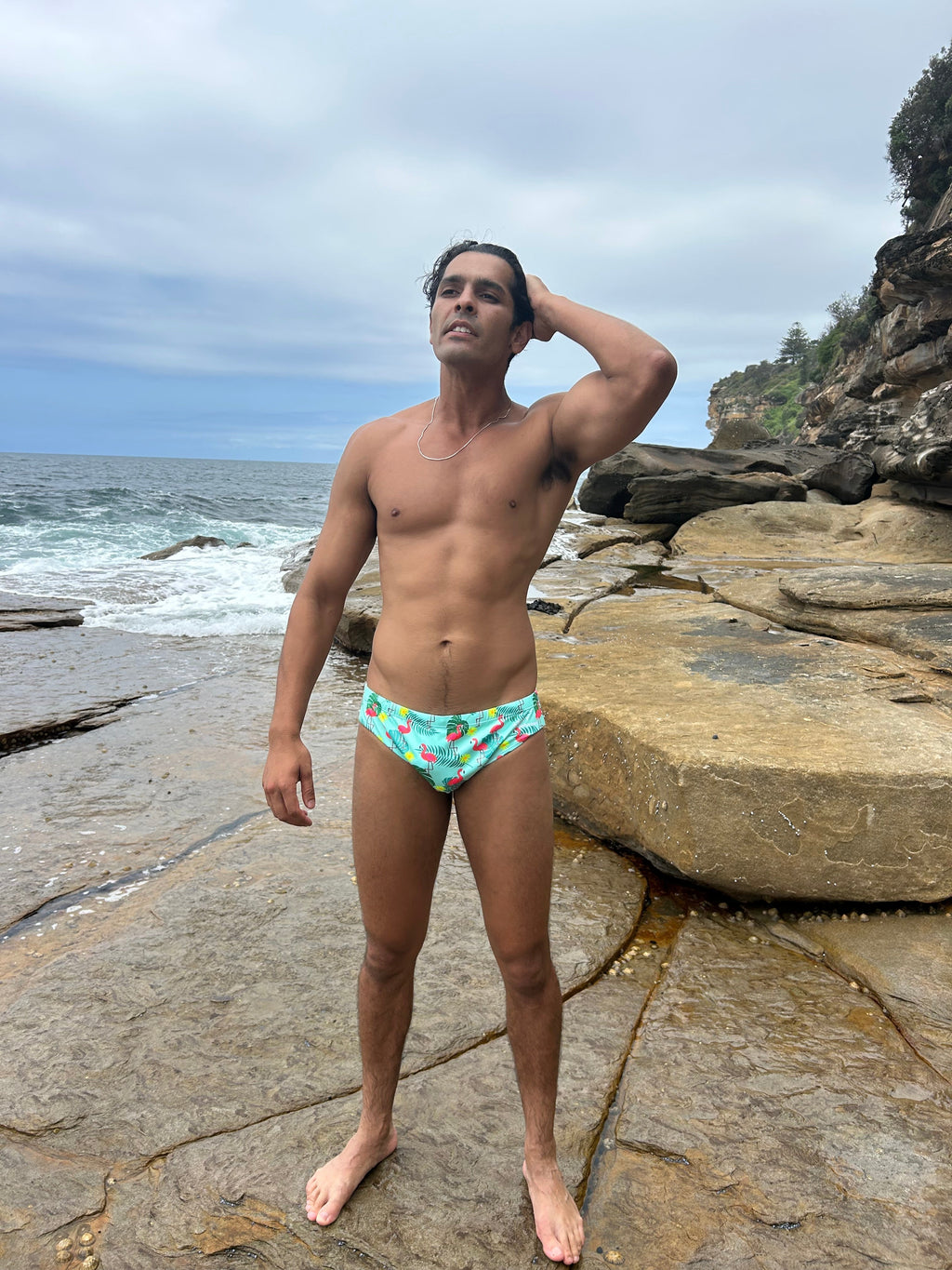 Male model wearing Sustainable Mens Swim Briefs with colorful pattern standing on rocky beach by ocean