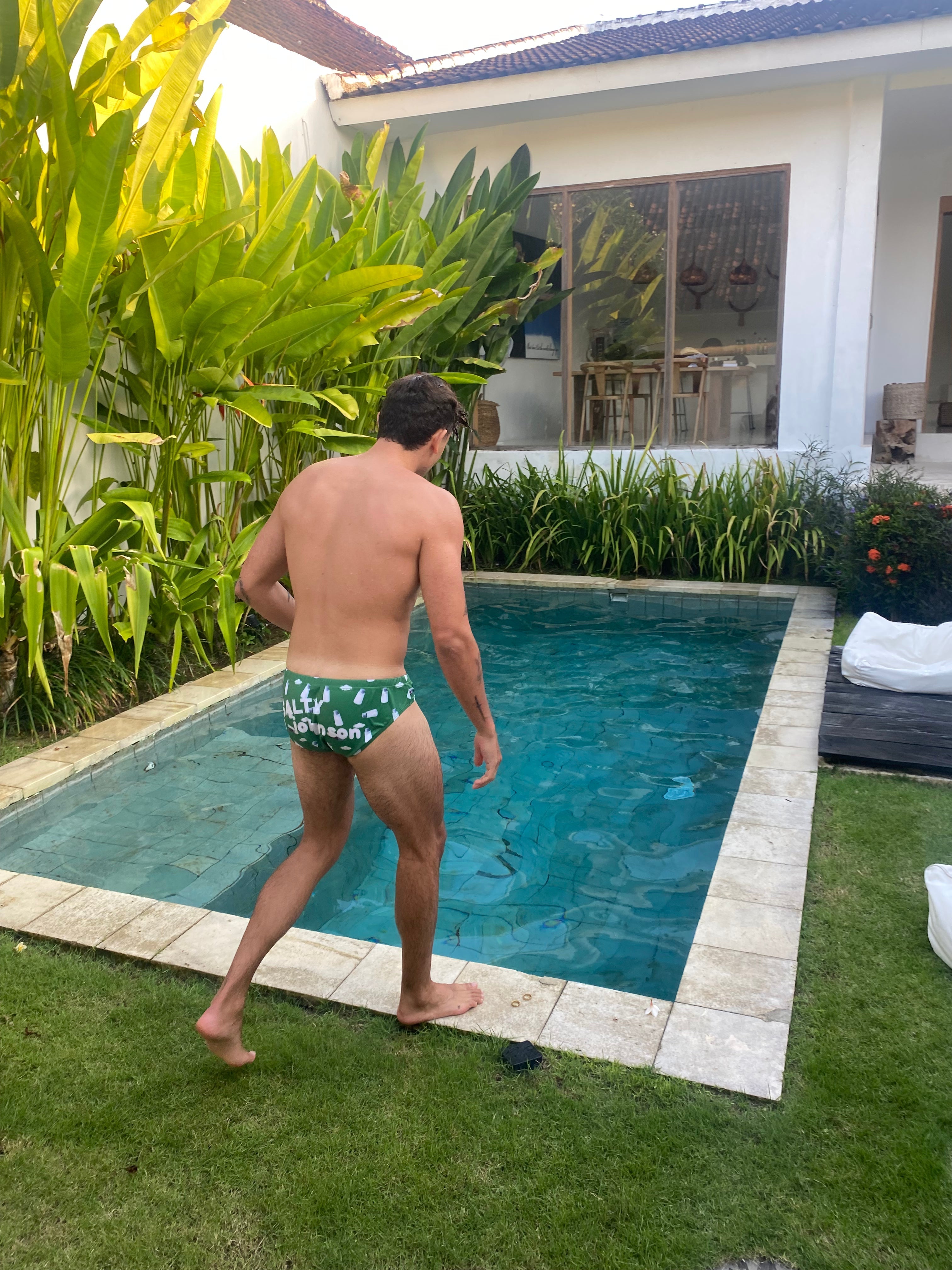 Man wearing sustainable mens swim briefs by Salty Johnson walking beside a backyard pool with tropical plants