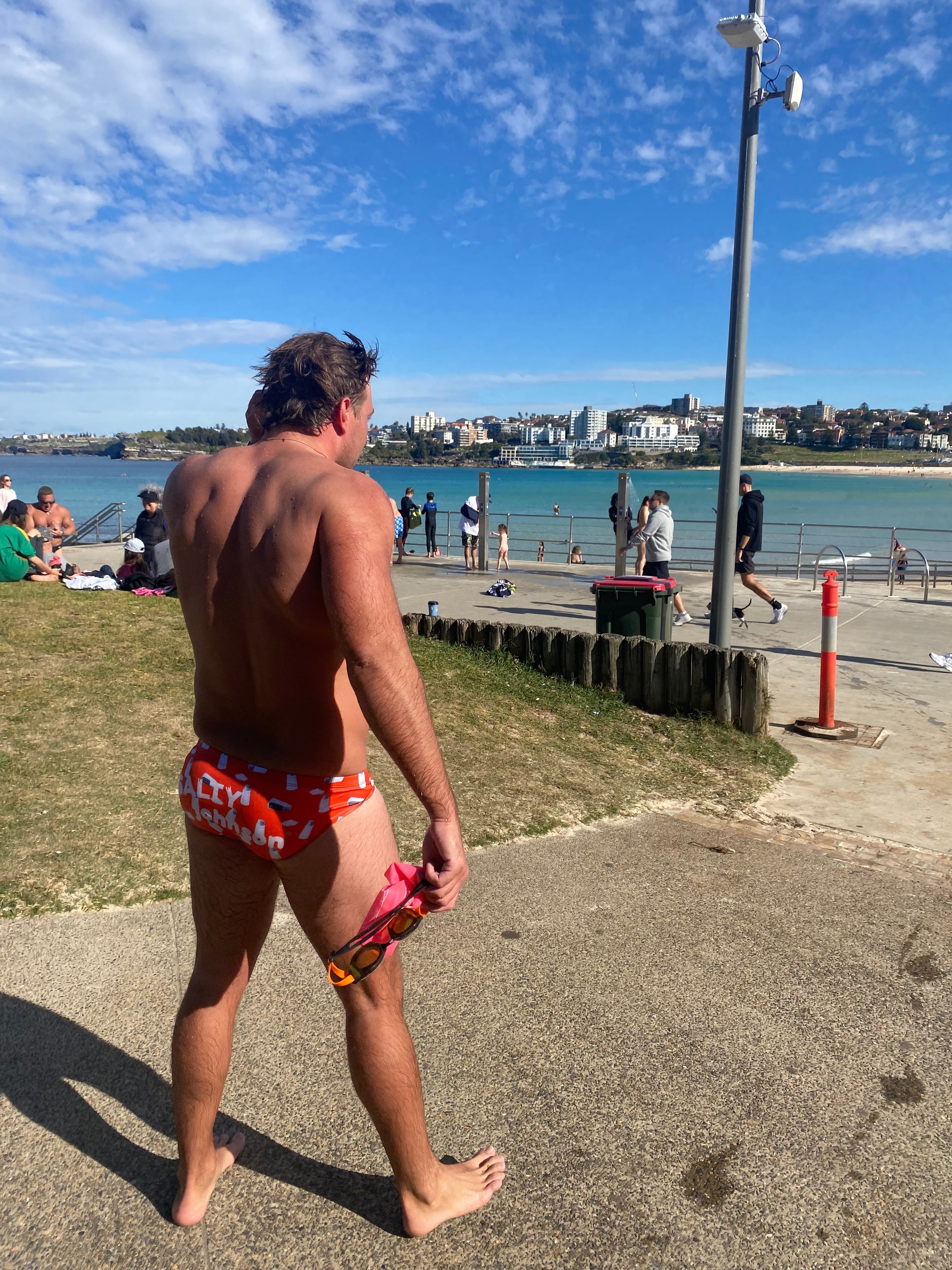 Man wearing Salty Johnson sustainable mens swim briefs in red with white text at a sunny beach park - Size 34