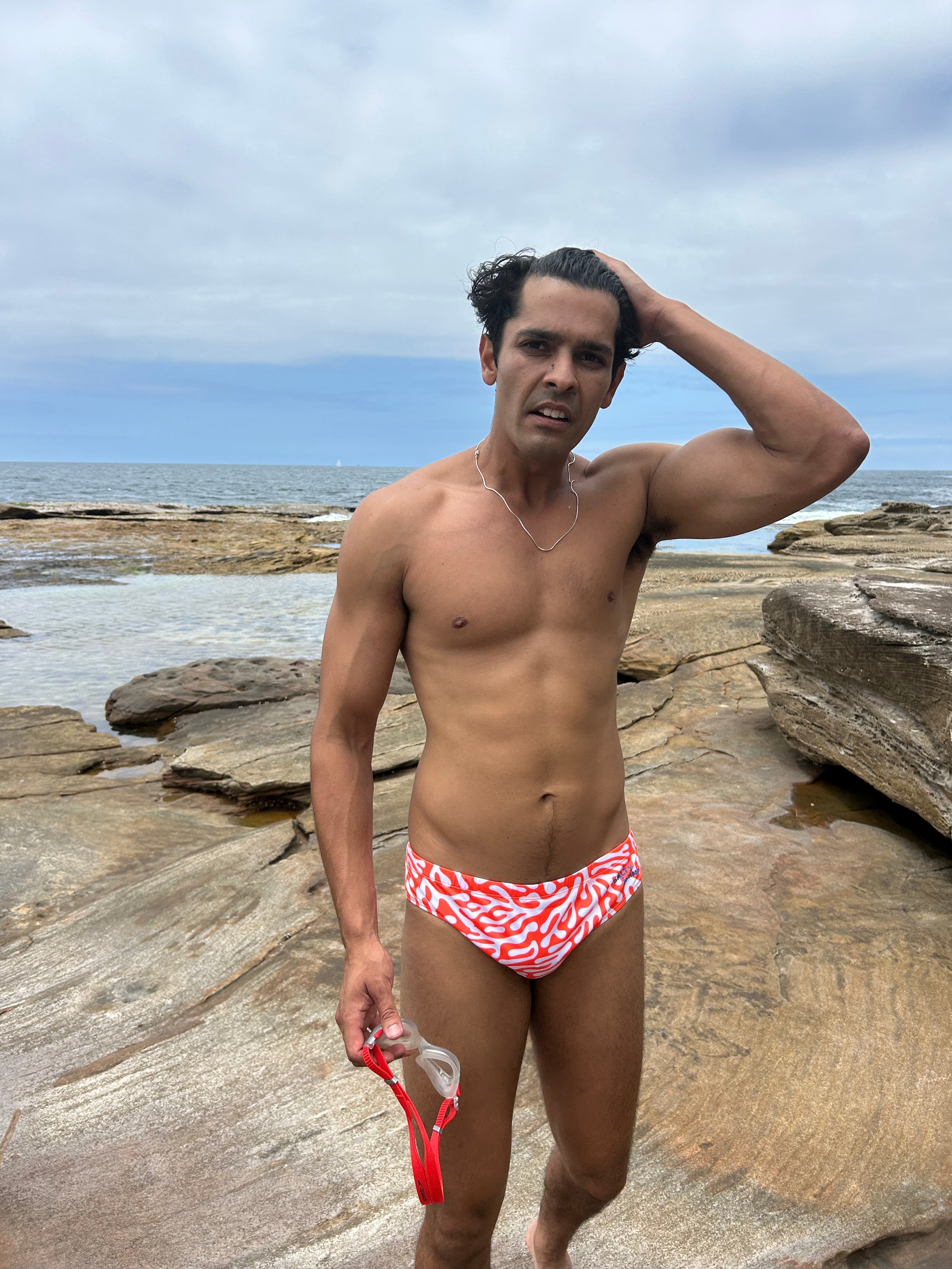 Sustainable Mens Swim Briefs by Salty Johnson AU in red and white worn by a man standing on rocky beach